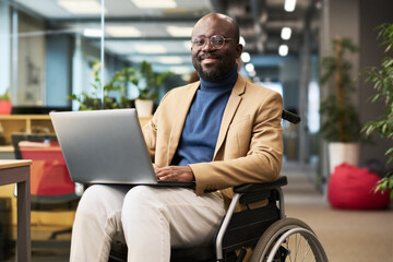 Happy young black man with disability sitting in wheelchair in office and looking at camera while analyzing online data on laptop screen