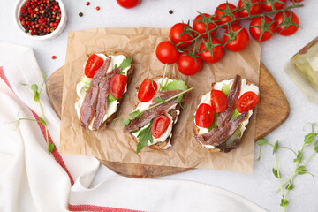 Delicious bruschettas with anchovies, tomatoes, microgreens and cream cheese on white table, flat lay