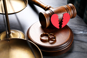 Divorce concept. Wooden gavel with wedding rings and broken red paper heart on black table, closeup