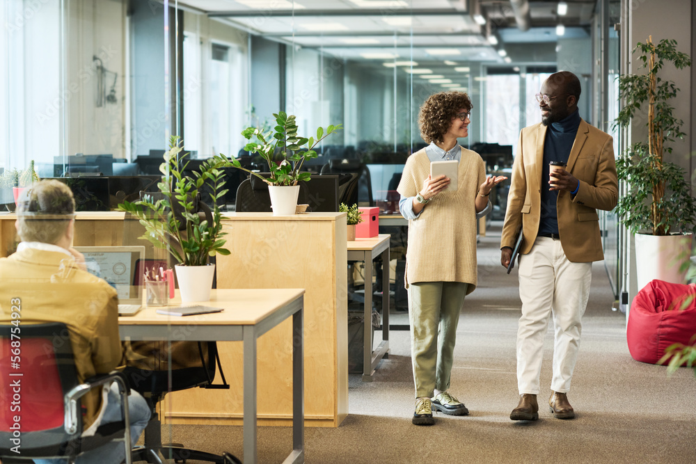 Poster Woman with tablet and her colleague with cup of coffee discussing main working points while moving along rows of desks in openspace office