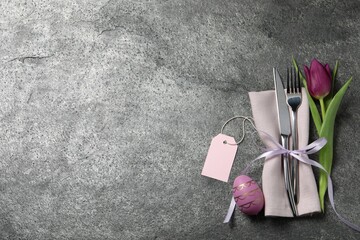 Cutlery set, painted egg and beautiful flower on grey table, flat lay with space for text. Easter celebration