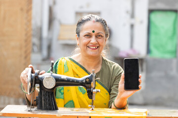Senior rural woman using sewing machine while showing smartphone with empty screen.