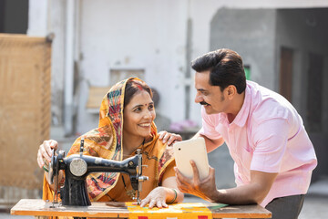 Indian rural woman using sewing machine and her husband showing digital tablet.