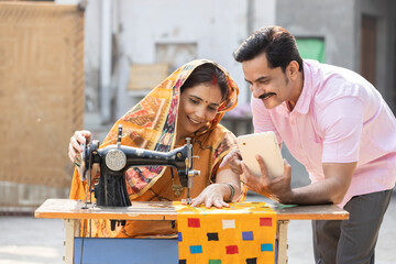 Indian rural woman using sewing machine and her husband showing digital tablet.