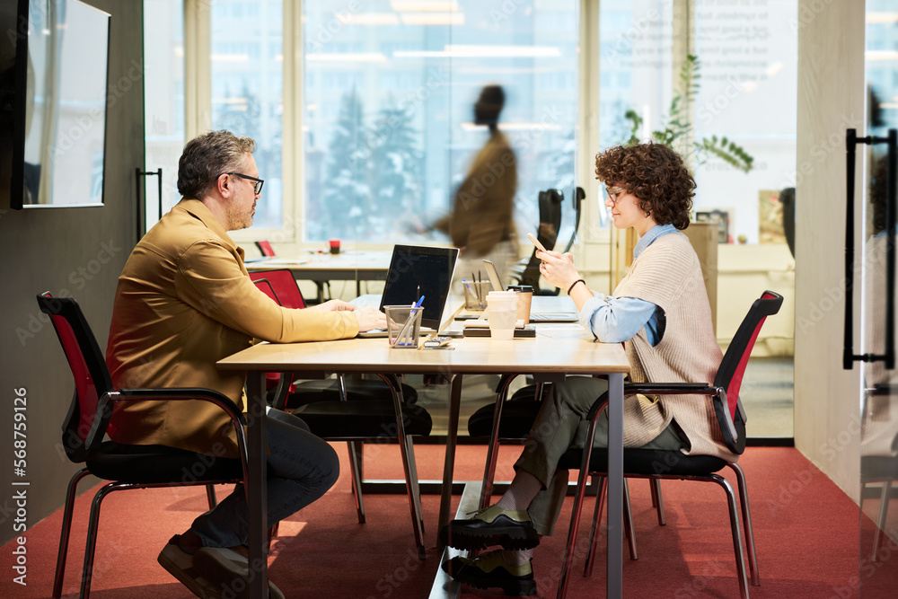Poster Side view of two coworkers sitting by table in openspace office and using laptop and smartphone against blurry outline of man passing by