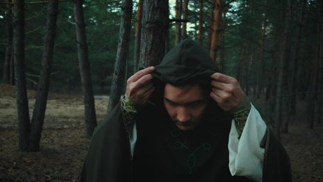 Portrait fantasy male elf prince takes off hood from head. Dark gothic dense forest. Mystical evil face fairy guy. Wizard man. Creative medieval clothes cloak cape green costume. Fashion model posing