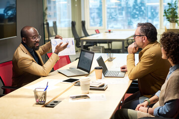Confident African American broker presenting graphic financial data to his colleagues during discussion of marketing development