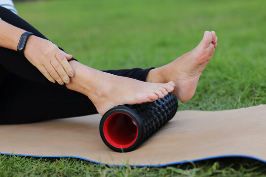 World health day. Unrecognizable woman warms up and works out the foot of the muscles of the body with a sport massage foam roller outdoors on green grass. Flexibility and mobility, body strength.