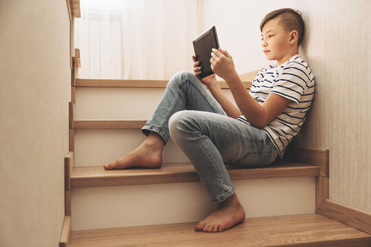 Teenage boy sitting on step of stairs using tablet playing video games on internet online at cozy home. Social media