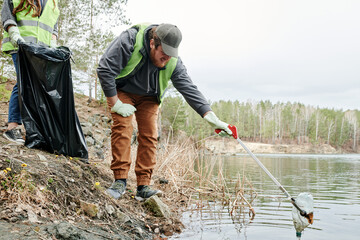 Man with picking stick reaching for dirty plastic bottle in lake