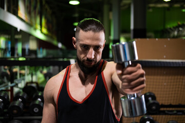 A handsome and brutal athlete with a beard holds dumbbells in his hands and does sports in the gym