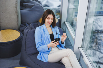 Business young woman in glasses with long dark hair in casual clothes smiling and looking at the phone, browsing the smartphone during a day off in the workspace