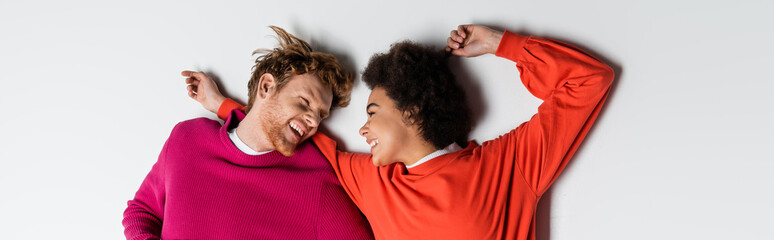 top view of happy redhead man and curly african american woman in magenta color clothes lying on white, banner.