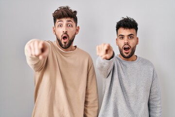 Young homosexual couple standing over white background pointing with finger surprised ahead, open mouth amazed expression, something on the front