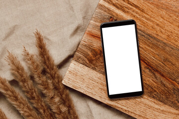 Blank mobile mockup screen on wooden desk with pampas grass on beige linen cloth. Mobile phone with...