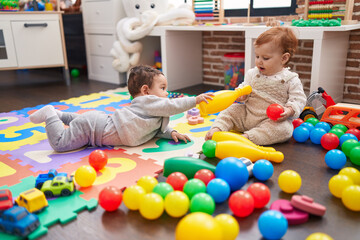 Two adorable babies playing with balls and bowling pin sitting on floor at kindergarten