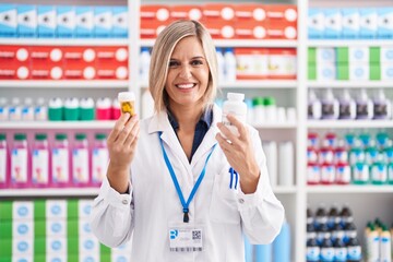 Young blonde woman pharmacist smiling confident holding pills bottles at pharmacy