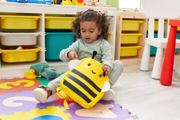Adorable hispanic girl student opening backpack sitting on floor at kindergarten