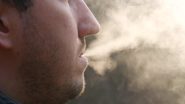 A handsome young man with a beard inhales the smoke of an electronic cigarette on the street close up view