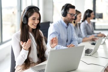 Smiling female call center operator in headphones with microphone consulting client on phone in customer support service while working with group of colleagues in row at bright office.