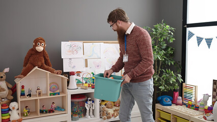 Young redhead man preschool teacher smiling confident collecting toys at kindergarten