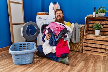 Redhead man with long beard putting dirty laundry into washing machine sticking tongue out happy with funny expression.