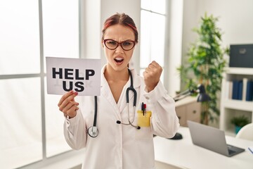 Young caucasian woman wearing doctor uniform holding help us banner annoyed and frustrated shouting with anger, yelling crazy with anger and hand raised