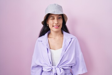 Young hispanic woman standing over pink background wearing hat winking looking at the camera with...