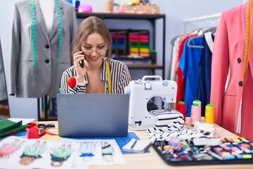 Fototapeta premium Young blonde woman tailor talking on smartphone using laptop at tailor shop