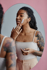 Candid young black woman with acne scars using face cream by mirror