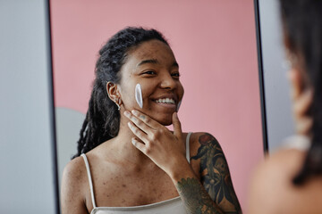 Candid smiling black woman with acne scars using face cream by mirror