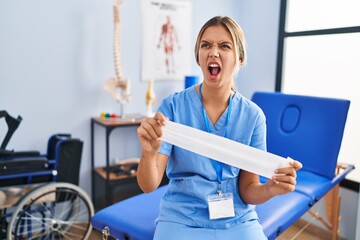 Fototapeta premium Young blonde woman holding therapy strip band angry and mad screaming frustrated and furious, shouting with anger. rage and aggressive concept.