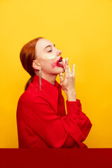 Beautiful young girl in red shirt with smudged red lipstick, licking whipped cream over yellow studio background. Food pop art photography. Complementary colors. Concept of art, beauty, food.