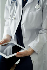 Unknown doctor woman using tablet computer or touchpad while standing near window in clinic. Dark blue work clothes of physician. Medicine concept