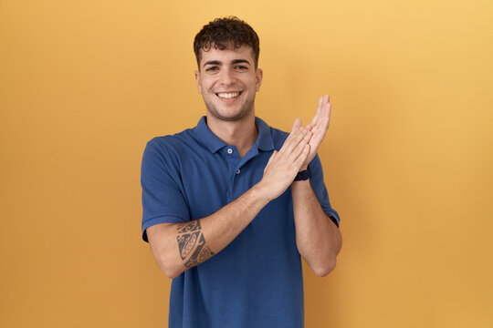 Young hispanic man standing over yellow background clapping and applauding happy and joyful, smiling proud hands together