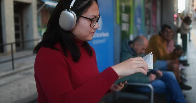 Young asian woman waiting at tram station while listening music playlist and reading book