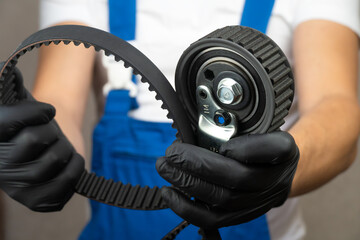 Auto mechanic in blue jumpsuit holds in his hands in black gloves roller belt tensioner of gas...