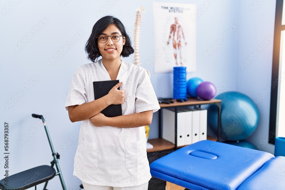 Sticker young latin woman wearing physiotherapist uniform hugging book at physiotherapy clinic