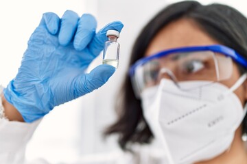 Young latin woman wearing scientist uniform and medical mask holding dose vaccine at laboratory