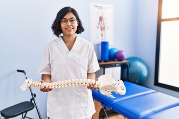 Young latin woman wearing physiotherapist uniform holding anatomical model of vertebral column at...