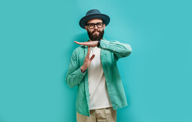 Young bearded hipster guy showing time out tired from work. Indoor studio shot against a blue...