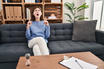 Young brunette woman at consultation office amazed and surprised looking up and pointing with fingers and raised arms.