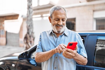 Senior grey-haired man using smartphone standing by car at street
