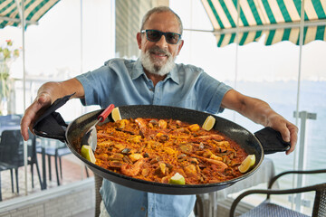 Senior grey-haired man smiling confident holding seafood paella at coffee shop terrace