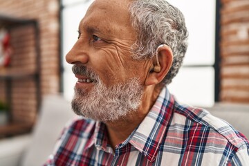 Senior grey-haired man using hearing aid sitting on sofa at home