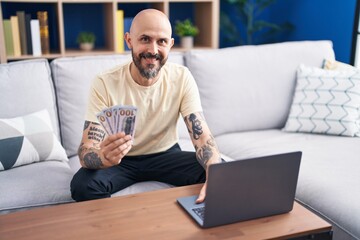 Young bald man using laptop holding dollars at home