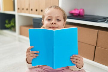 Adorable blonde girl student reading book sitting on floor at library school