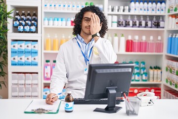 Hispanic man with curly hair working at pharmacy drugstore covering one eye with hand, confident smile on face and surprise emotion.