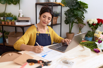 Middle age hispanic woman florist using laptop writing on document at flower shop