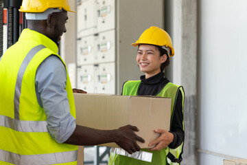 Young Asian woman and black man worker in safety vest and yellow helmet holding cardboard box preparing product for shipment at warehouse factory